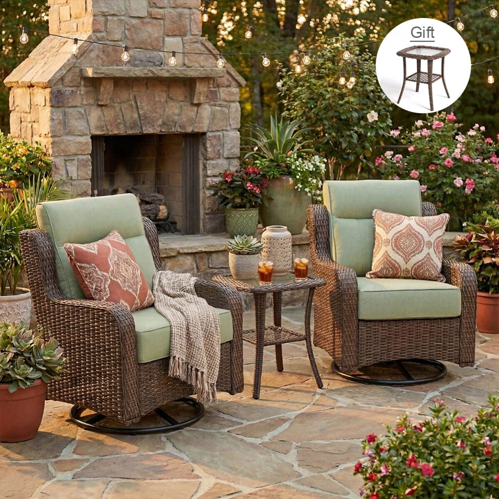 Two brown wicker patio swivel armchairs with light sage green cushions and patterned throw pillows, set on a stone patio in front of a stone fireplace. A small round dark wicker side table is between them, with a throw blanket draped over one chair.