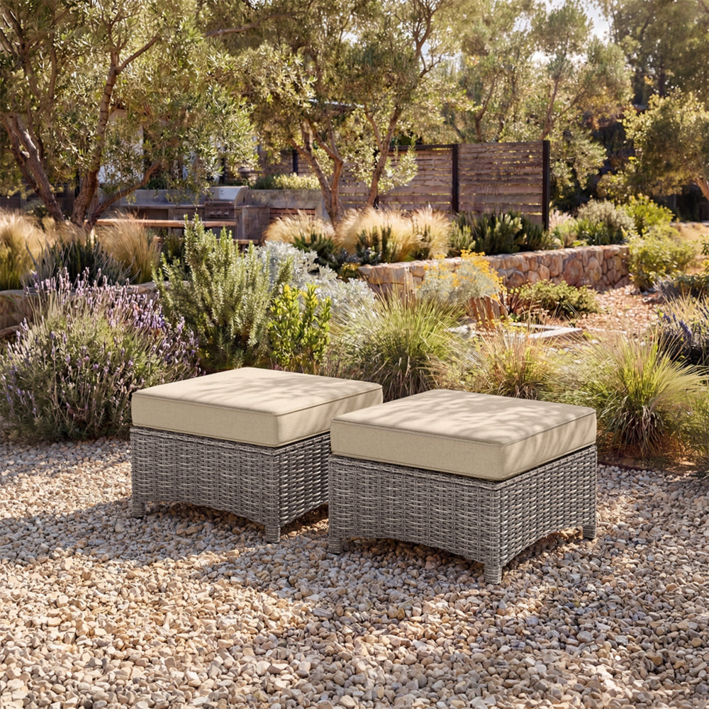 Two wicker ottomans with beige cushions on a gravel surface in a garden setting.