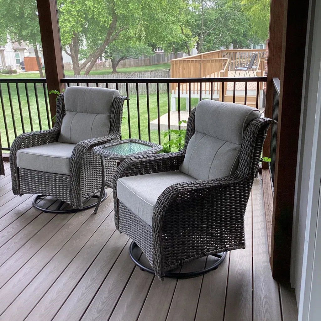 Two dark gray wicker patio swivel chairs with light gray cushions and a small glass-top side table on a wooden deck, with a backyard and trees visible in the background.