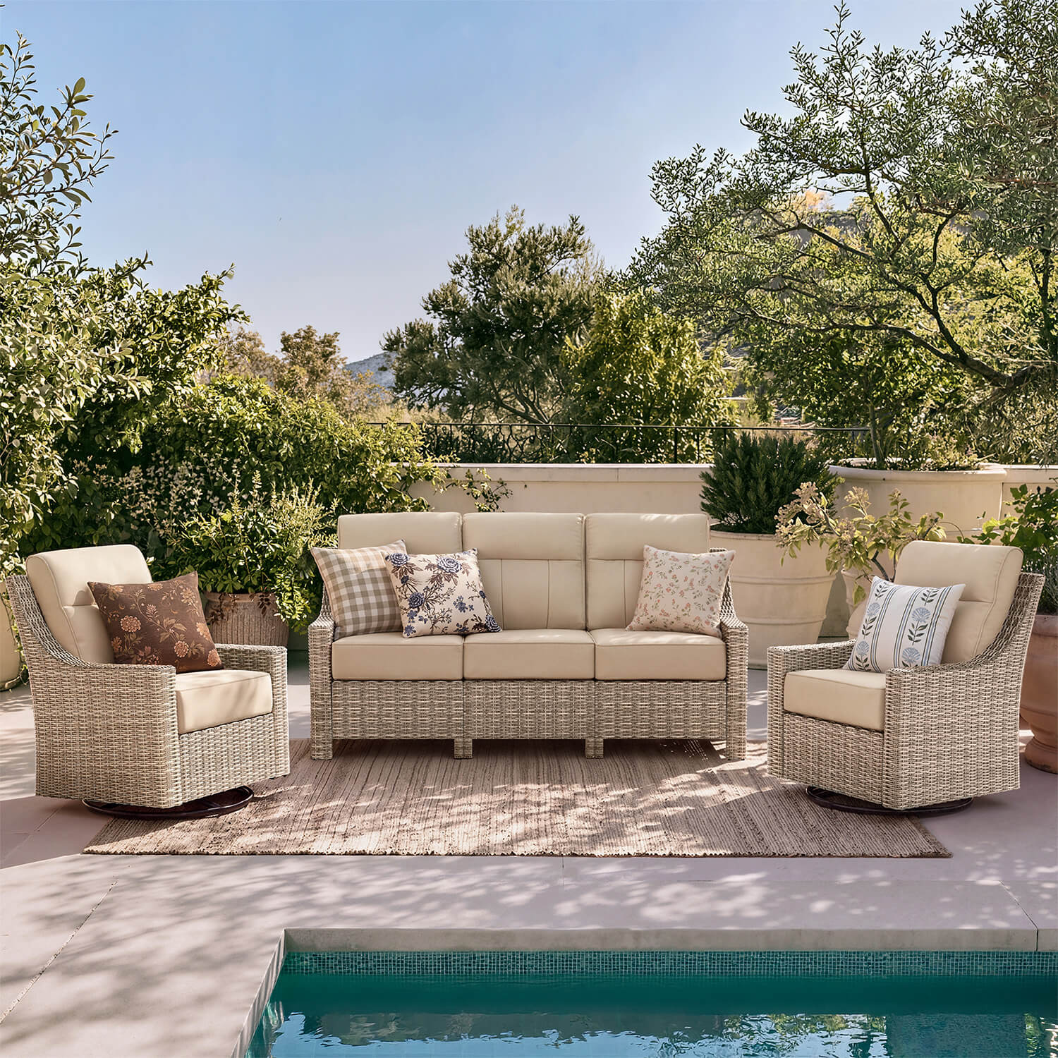 Outdoor patio furniture set including a three-seat sofa, two armchairs, and a woven rug on a poolside terrace with greenery and blue sky in the background.