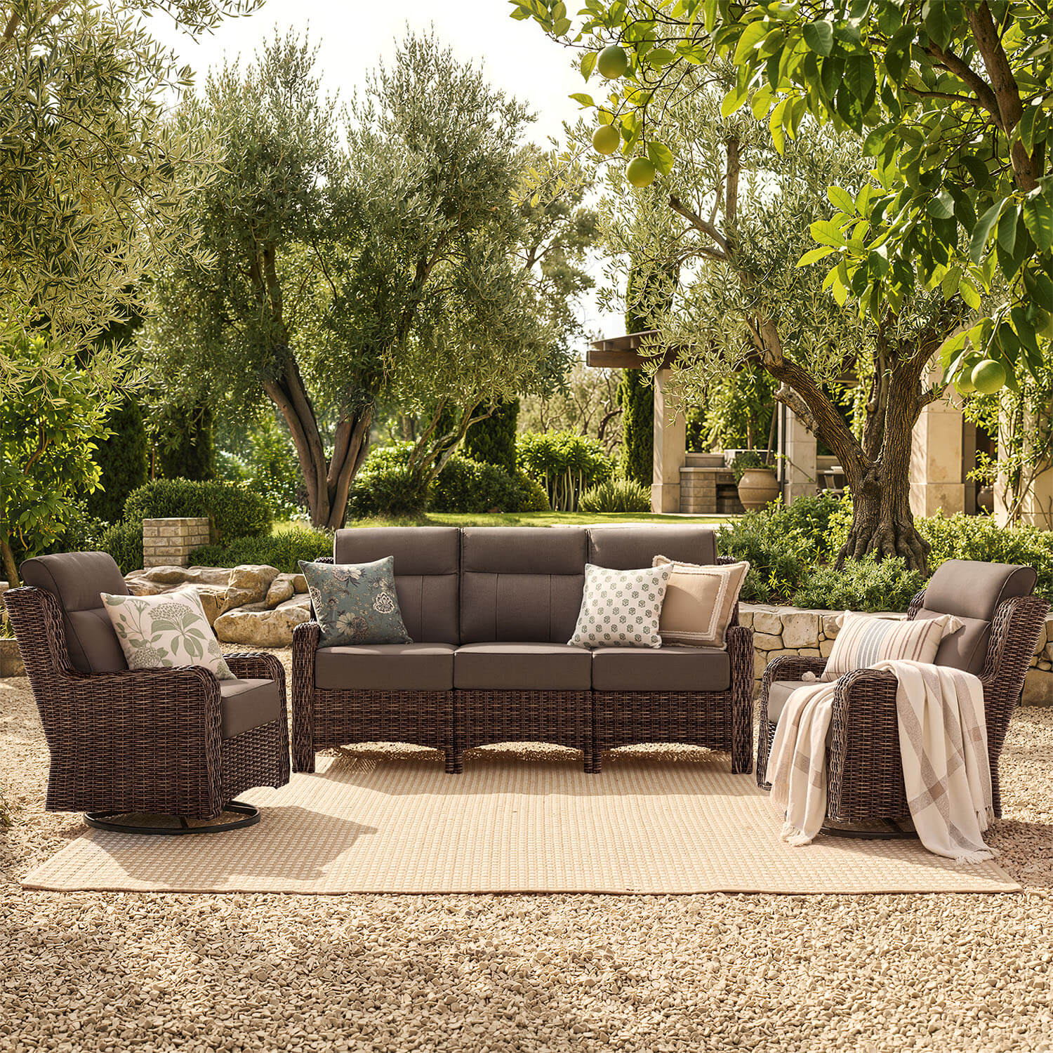A 5-piece outdoor patio furniture set including a 3-seat sofa, 2 armchairs, and a matching coffee table, all in dark brown wicker with dark brown cushions, styled on a beige woven rug in a garden setting.