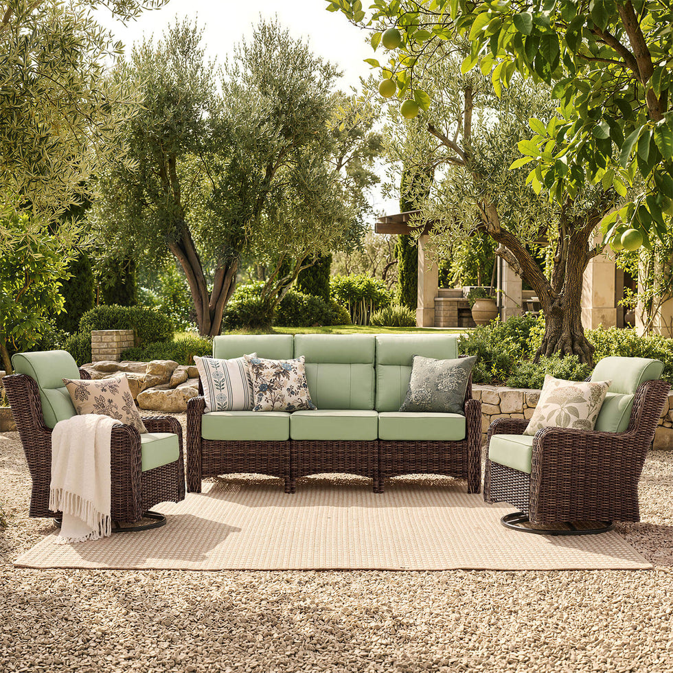 Outdoor patio furniture set with a three-seat sofa, two armchairs, and patterned throw pillows in a garden setting. The furniture features dark brown woven wicker frames and light sage green cushions, arranged on a beige outdoor rug over a gravel surface.